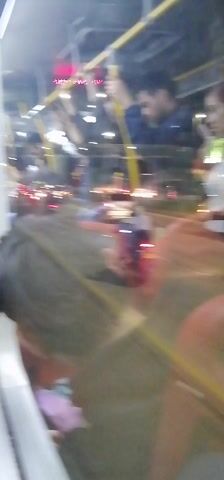 The video appears to be shot from inside a moving bus at night. The camera's view is directed towards a side window, showing the street and the bus's interior. Several people are standing and holding onto the bus's handrails. Outside, there are cars and motorcycles on the road, identifiable by their headlights and taillights. The street is illuminated by city lights, creating reflections on the bus window. The video is somewhat blurry due to the motion and the dim lighting conditions.