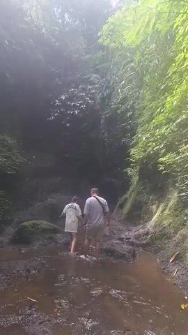 Two people are walking through a shallow stream in a narrow gorge. The person in front is wearing a white shirt and dark shorts, and the person behind them is wearing a grey t-shirt and light-colored shorts. The gorge is lined with lush green vegetation, and the ground is muddy and rocky. Sunlight filters down from above, creating a misty effect.