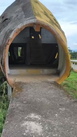 The video shows a wide-angle, eye-level shot of a large, dilapidated concrete structure shaped like a fish head. The entrance to the fish head is wide and cavernous, with a concrete pathway leading up to it. Inside the fish head, there are visible stairs leading upwards, suggesting multiple levels or rooms within the structure. The exterior of the fish head is painted yellow and orange, but the paint is peeling and worn, indicating age and neglect. The environment appears to be outdoors, with greenery visible to the sides and in the background, suggesting a natural or overgrown setting. The sky above is overcast. The camera is stationary and does not capture any people or movement.