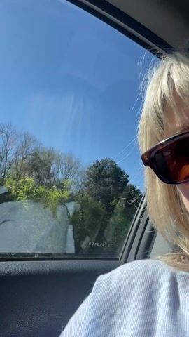 A woman with short blonde hair is sitting in the passenger seat of a car. She is wearing sunglasses and a light blue top. The car is moving, and a wind turbine can be seen through the window. The sky is blue and sunny.