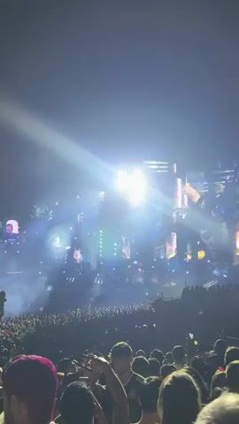 The video displays a crowded outdoor music festival at night. A large audience is visible in the foreground, silhouetted against a stage filled with elaborate structures and bright lights. Beams of white and blue light emanate from the stage, illuminating the fog and smoke that fill the air. The stage itself features tall, futuristic-looking buildings adorned with lights. The crowd appears to be enjoying the performance, with many people holding up their phones to record. The overall atmosphere is energetic and festive.