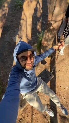 This video captures a person swinging on a swing set in a park-like setting. The subject, wearing a blue hoodie, a baseball cap, and sunglasses, is holding onto the chains of the swing. The background reveals tall trees, suggesting a natural environment, likely during daylight hours given the strong sunlight and shadows. The weather appears clear and sunny. Other people are visible in the background, but they are out of focus. The camera angle is low, looking up at the swinging person and the trees. The overall mood seems to be one of enjoyment and recreation. There is no visible text in the video. The location context provided is Онохино, Russia.