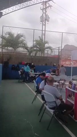 The video is a handheld vertical shot of an outdoor event. It captures people in casual attire seated on white folding chairs, engaged in conversations and relaxation. Some individuals are seen walking around in the background. A red booth with products displayed on shelves is visible on the right side of the frame. In the background, there is a chain-link fence, trees, and a mountain range under an overcast sky. A man wearing a blue shirt and a baseball cap briefly looks at the camera. The video captures the atmosphere of a community gathering or a casual outdoor event.