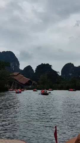 The video shows a river scene with several small rowboats filled with people wearing orange life vests. They are scattered across the water, some rowing, with others possibly just observing the scenery. In the background, there are lush green mountains and a structure with a thatched roof. The sky is overcast and grey. The camera appears to be positioned on another boat, with a glimpse of an orange oar visible in the lower right corner of the frame. The overall mood is tranquil and scenic.