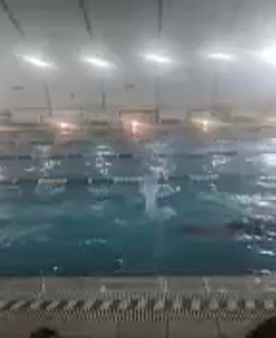 This is a video of an indoor swimming pool. The pool has lanes marked by ropes and buoys. The water is a clear blue color, and there are lights overhead. The pool deck is visible at the bottom of the frame, with a textured surface. The video is slightly blurry and grainy, suggesting it was filmed in low light conditions. The water appears to be rippling, indicating movement.