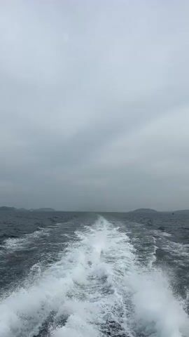 The video shows the view from the back of a boat moving through the ocean. The water is dark and choppy, and the boat is leaving a white foamy wake. In the distance, there are islands or landmasses on the horizon. The sky is overcast with gray clouds. There are no people visible in the frame.