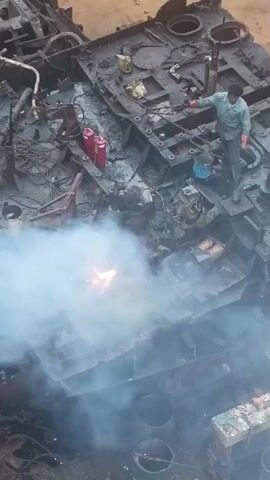 The video shows an aerial view of a man working on a large, heavily damaged metal structure. The structure appears to be a vessel or a large piece of machinery, with visible signs of wear and tear, including rust and missing components. The man is dressed in work clothes and gloves and is engaged in some sort of cutting or welding activity, indicated by the bright sparks and smoke emanating from the metal. He is positioned on top of the structure, near some red canisters and a blue bucket. The overall scene suggests a demolition or dismantling process. The setting appears to be an industrial area, possibly a shipyard or a scrap yard, given the presence of various metal components and the general state of disrepair. The lighting suggests it is daytime, but no specific time or weather conditions can be determined. There is no visible text in the frame. The activity is focused on the disassembly of the large metal object, with the worker actively involved in the process.