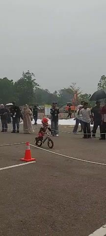 A group of people are gathered outdoors near a road, possibly at a race event. There are orange traffic cones and white ropes on the asphalt. In the foreground, a child in racing gear is on a small bicycle, appearing to be in motion. Further back, spectators are observing. Some individuals are holding umbrellas. In the background, there are trees, hills, buildings, and parked vehicles, including an ambulance. The sky is overcast.