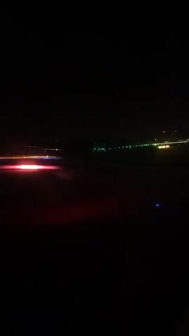 The video shows a view from an airplane window at night. The runway lights are visible in the distance, appearing as colorful dots of red, blue, green, and yellow against the dark sky. The lights are in a line, indicating a runway. There are no people or other objects clearly visible in the frame. The overall impression is one of takeoff or landing at night.