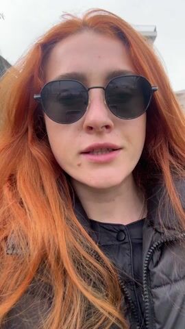 A woman with bright red hair and black sunglasses is walking outside on what appears to be a cloudy day. She is wearing a black jacket. Buildings and a crane can be seen in the background. The camera moves as she walks, and at times, her face fills much of the frame. She is singing or speaking.