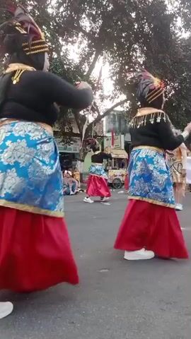 A group of dancers in traditional attire perform a street dance. The dancers wear black tops, blue patterned skirts, and red skirts. They also have tall, feathered headdresses. Some of the dancers are facing away from the camera, while others are partially visible from the side. The street is paved, and there are trees and buildings in the background. Spectators are seated along the side of the street, watching the performance. The overall atmosphere appears to be festive and lively.