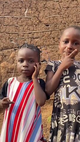 Two young Black girls stand in front of a textured, earthy-brown wall that appears to be made of mud or clay, possibly a dwelling in Ufu Esioni, Nigeria. The girl on the left, with her hair styled with twigs and blue string, wears a dress with vertical stripes in red, white, and blue. She has her left hand touching her chin and her right hand raised with her index finger pointing outwards. The girl on the right wears a black and white dress with a repeating pattern of text that appears to be brand logos, including "GUCCI". She holds her right hand up, forming a peace sign with her index and middle fingers. Both girls have neutral or serious expressions. The lighting suggests it is daytime, possibly late afternoon, given the warm tones of the wall. The scene evokes a sense of everyday life in a rural setting.