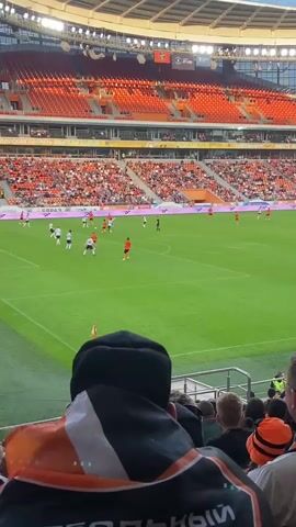 The video shows a soccer game from the perspective of a spectator. The spectator is wearing a black hat and an orange and black scarf. The stadium is filled with spectators, many of whom are wearing orange. The field is green, and the players are wearing white and orange uniforms. The game appears to be in progress, with players on both sides of the field. The lighting suggests it is either daytime or the stadium lights are on. The focus is on the field of play, with the stands and other spectators blurred in the background.