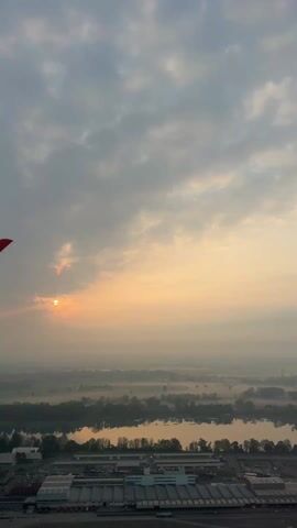 The video presents a scenic, elevated view of a landscape at sunrise or sunset. The sun is visible through a light cloud cover, creating a soft, diffused light. Mist hangs over the fields and water bodies, partially obscuring the view. A large industrial or commercial complex is visible in the foreground, complete with parking lots, buildings, and vehicles. A small red object is visible in the top left corner of the frame. The sky is filled with varied cloud formations, with a range of grey and pastel colors from the setting sun.