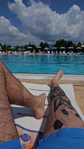 The video shows a first-person perspective of someone lying on a lounge chair by a swimming pool. Their legs are visible in the foreground, one with a tattoo of a panda and other designs, the other with more hair. The pool is filled with blue water and lined with lounge chairs and umbrellas. Several people are seen swimming and relaxing in and around the pool. The sky is blue with scattered white clouds. The environment appears to be a sunny day at an outdoor water park or resort.