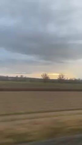 The video shows a view from a moving vehicle, likely a train or car, across a rural landscape. The foreground is a field, possibly recently plowed, with brown soil. Beyond that is a narrow strip of green vegetation, followed by another field. In the distance, there are a few bare trees silhouetted against a bright horizon, suggesting either sunrise or sunset. The sky is mostly covered with grey clouds.
