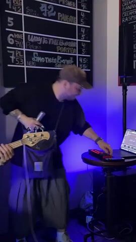 The video shows a man with a beard and a baseball cap, dressed in a black shirt and oversized gray pants. He is holding a microphone in his right hand and a bass guitar slung over his shoulder. He is looking towards the camera and smiling, making gestures as if he is singing. Beside him, to the left, another man with a dark baseball cap is playing the bass guitar. Behind them, there's a black menu board with handwritten items and prices in white chalk. To the right of the first man, a small round table holds a laptop and some electronic gear, with blue lights illuminating the scene. The background also includes a brick wall and posters, suggesting they might be performing in a bar or a similar venue.