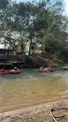 The video shows a river with several people tubing down the water.  The people are on red tubes and seem to be relaxing and enjoying themselves. The river has a sandy bank on the left side.  Behind the bank there is a green lawn with trees. There is some construction, and what appears to be a small building on the lawn. The sun is shining, and the sky is blue.