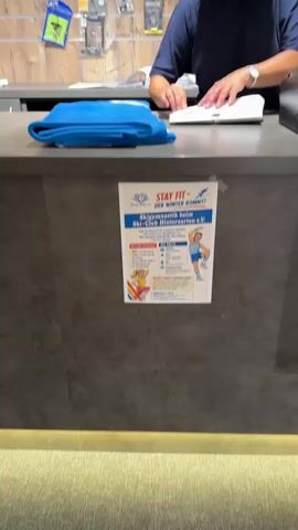 The video shows a close-up view of a counter in what appears to be a reception area or shop. A person, partially visible from the chest down, is standing behind the counter. They are wearing a dark blue shirt and have their hands on a white object, possibly a book or a notepad, and appear to be handling papers. In the foreground, on the counter, is a folded blue towel. A flyer with the title "STAY FIT - DER WINTER KOMMT!" is attached to the front of the counter. The flyer features graphics of two cartoon characters, a male and a female, and text in German related to a ski club and gymnastics. The background shows shelves with various items, including what looks like sports equipment or accessories. The lighting is moderate, and the overall environment seems to be indoors.