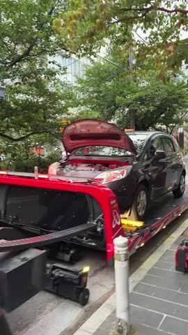 A black car with its hood open is being loaded onto a red flatbed tow truck from the front. The car is partially on the flatbed, and its front wheels are secured with yellow chocks. The tow truck has a red cab and a black flatbed with a textured surface for traction. The scene takes place outdoors on a paved surface next to a sidewalk. Trees and buildings are visible in the background. The sky is overcast, suggesting it might be an early morning, late afternoon, or cloudy day.