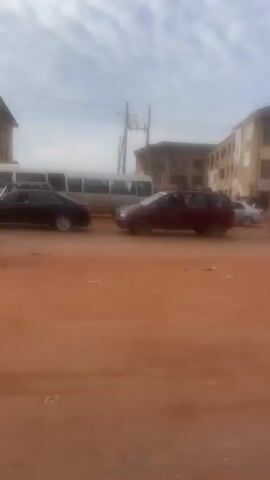 A daytime video shows a static, eye-level view of a street with a reddish-brown dirt or sand ground. Buildings with multiple stories and various signs, including "EMANDO PLAZA" and "LG," line the background. Utility poles and wires are visible against a partly cloudy sky. Numerous vehicles, including cars, SUVs, a bus, and a small white truck, drive or are parked on the street. At 0:01, two men walk from left to right across the foreground; one wears a yellow reflective vest and carries a long, dark object, while the other wears a red beanie and a green jacket and looks at a phone.
