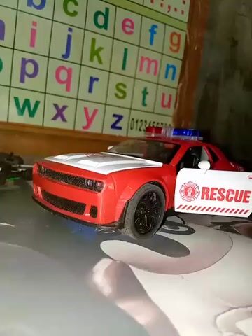 The video shows a red and white toy rescue vehicle with its driver's side door open. The vehicle has blue and red lights on its roof. The word "RESCUE" is visible in large white letters on the open door. The background is out of focus but appears to be a colorful chart with letters of the alphabet and numbers. The toy car is positioned on a surface that has a faint circular pattern.
