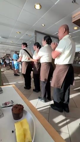 Four individuals, likely staff, are clapping in unison while facing away from the camera. They are wearing matching uniforms consisting of white shirts, brown aprons, and dark pants. The setting appears to be an indoor dining area, possibly a buffet, with a long table in the foreground. On the table, there is a plate with a partially eaten pastry, a cupcake, and slices of pineapple. In the background, other people are visible, some standing in line. The lighting suggests it is daytime.