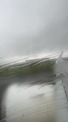 This is a first-person view from an airplane window. The view is slightly obscured by water droplets on the window. An airplane engine is visible on the right side of the frame. The plane appears to be on a runway, as indicated by the asphalt surface and the grass bordering it, and it is moving, suggesting takeoff or landing. The sky is overcast, and the lighting suggests it is daytime. The plane is moving from left to right within the frame as it gains speed.