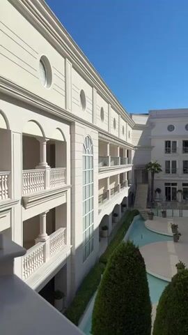 The video shows a courtyard of a hotel or resort with a clear blue sky. The building is white and neoclassical in style, with columns, balconies, and arched windows. There is a narrow, shallow pool or water feature in the center of the courtyard, surrounded by landscaped bushes and trees. The camera pans slowly across the courtyard, offering a wide view of the architecture and the water feature. No people are visible in the video.
