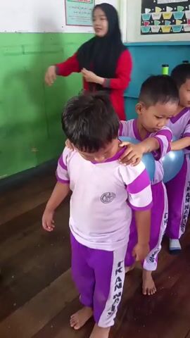 Children in purple and white uniforms are playing a game with blue balloons in a classroom. They are lined up and holding the balloons between their knees or thighs as they walk. The classroom has colorful decorations on the walls, including what appear to be drawings of clouds and stars. There are also some shelves and a cabinet in the background. The children are smiling and engaged in the activity, suggesting a fun and playful atmosphere.