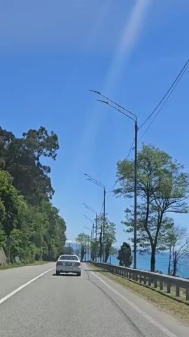 The video captures a scenic drive along a coastal road in Primorskoe, Abkhazia, on a bright, sunny day. The perspective is from inside a moving vehicle, looking forward and slightly to the right.

The scene features an asphalt road with a white dashed line on the left. A metal guardrail separates the road from a grassy bank that slopes down towards the sea on the right. The calm, turquoise-blue sea stretches to the horizon under a clear, brilliant blue sky, with some visible lens flares or wispy clouds in the upper left.

Tall, leafy green trees line the roadside, intermittently framing and partially obscuring the view of the water. Modern streetlights with double arms extend over the road, and power lines are visible running along poles. A white car can be seen driving ahead on the road. Brief moments show prominent orange and yellow reflections on the car window, likely from sun glare or interior objects, partially obscuring the view of the passing trees and sea. A tall, dark metal pylon or tower is also visible through the trees in some shots.

The overall impression is one of pleasant travel along a picturesque seaside route. No explicit text is visible in the video frames.