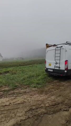 A yellow excavator is digging in a muddy field on a foggy day. A white van is parked to the right of the excavator, facing away from the camera. The excavator's arm is extended and its bucket is in the dirt. The van has a ladder on its roof. There is a house in the background on the left.