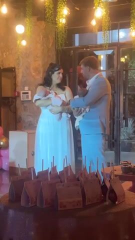 A man and a woman are standing at a table covered with a sparkly red tablecloth. They are both holding babies. The woman is wearing a white dress and the man is wearing a blue suit. There are many small, triangular paper bags on the table in front of them. The room is decorated with hanging plants and lights.