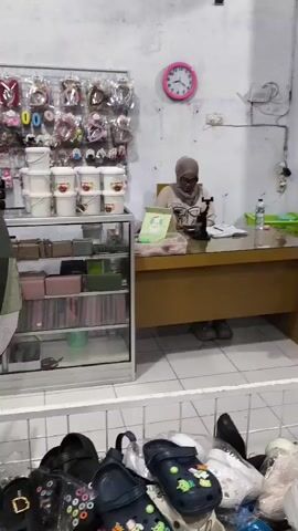 The video opens with a man standing and holding a black bag and a gold frame. He is facing a counter where a woman wearing a hijab is seated. Behind the man, there are shelves stocked with various items, including hair accessories and containers. To the right of the counter, there is a pink shelving unit filled with more items. In the foreground, there is a display of footwear. The man turns towards the seated woman, presenting the items he is holding. The woman looks at him and appears to be interacting with him. The man then places the items on the counter, and the woman leans forward, seemingly examining them. The scene takes place in what appears to be a retail store.