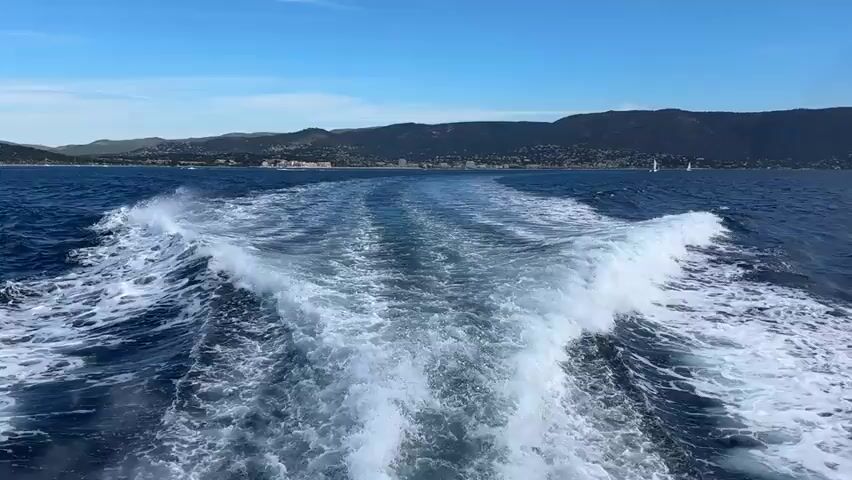 The video shows a view from the back of a boat moving through the water. A prominent wake trails behind the boat, creating white foam and turbulent waves. The water is dark blue. In the distance, a coastline is visible with hills and a town or city along the shore. The sky is clear and blue with a few wispy clouds. Several sailboats are also visible on the water in the distance. The overall impression is of a sunny day at sea.