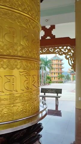 The video shows a large, golden prayer wheel with intricate carvings in a temple. The wheel is stationary and takes up the left side of the frame. Through an elaborately decorated doorway, a multi-tiered pagoda is visible in the distance. There is a bench nearby in the courtyard. The floor inside the temple is made of polished tiles, reflecting light. The scene appears to be peaceful and still.