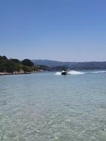 A jet ski is being ridden in the ocean on a sunny day. The water is clear and blue, and the sky is a bright blue. There is a sandy beach in the background, with trees and some buildings. The jet ski is creating a spray of water as it moves across the ocean.