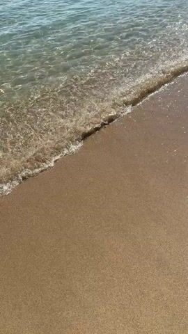 A person's feet are visible as they stand on a sandy beach with the ocean waves gently lapping at the shore. The water is clear, revealing the sandy bottom beneath. In the background, the ocean extends to the horizon under a clear blue sky. Rocky formations are visible along the coastline in the distance. The sun is shining, creating a bright and serene atmosphere. The video focuses on the interaction between the water and the sand, capturing the tranquil beauty of the beach.