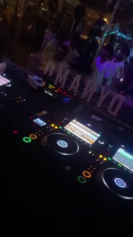 The video shows a DJ setup in a dimly lit, crowded outdoor venue. The DJ equipment, including turntables and a mixer, is in the foreground. In the background, a crowd of people are visible, some appearing to be dancing or socializing. The atmosphere suggests a nightclub or bar setting, with ambient lighting and music likely playing. The word "DYNAMO" is visible in large lettering behind the DJ.