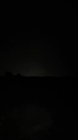 A dark, outdoor scene. A faint light source illuminates a flat, open area with some low vegetation. In the distance, silhouetted hills or mountains form a dark horizon against a slightly lighter sky. A group of several indistinct figures are visible in the foreground, scattered across the illuminated area. The lighting suggests it is nighttime or very low light conditions.