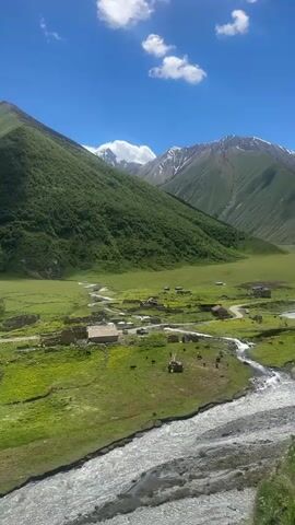 A panoramic aerial view of a lush green valley with a winding river. In the middle ground, a small village is nestled, featuring stone buildings and some grazing livestock. The surrounding mountains are steep and covered in vegetation, with snow-capped peaks visible in the distance. The sky is a vibrant blue with scattered white clouds. The camera is static, offering a fixed perspective of the landscape.