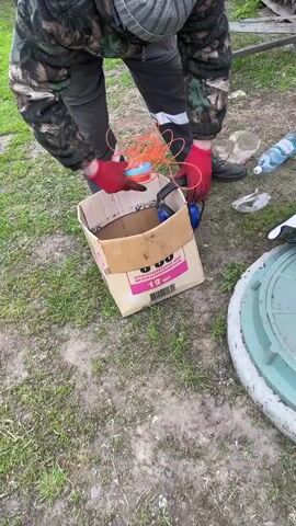 A man wearing a camouflage jacket, grey trousers, a grey and white striped beanie, black shoes, and red gloves is bending over a cardboard box. The box is filled with orange trimmer line and a set of blue earmuffs. The man is taking a white plastic container out of the box. Next to the box, on the grass, are plastic bottles and a white object wrapped in a black and white cloth. To the right, there's a round concrete manhole cover. The ground is covered in grass and patches of dirt. A wooden structure is visible in the background.