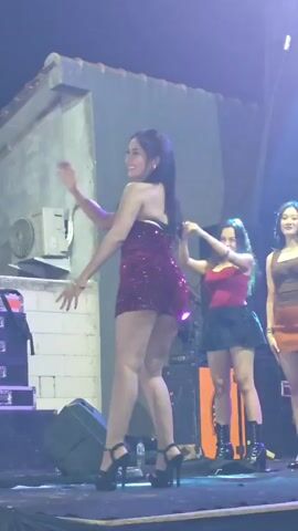 A group of young women are on a stage at night. The woman in the foreground is wearing a sparkly red dress and high heels. She is dancing and smiling. Another woman in a red top and black skirt is standing behind her, also smiling. A third woman in a brown top and orange skirt is visible on the right. The stage has speakers and lighting equipment. The background appears to be a building with some outdoor air conditioning units.