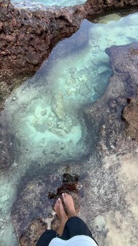 The video shows a person standing barefoot on rocky ground next to a tide pool. The water in the tide pool is clear and a light turquoise color, revealing rocks and sand at the bottom. The pool is surrounded by dark, textured rocks that vary in color from brown to grey. The person is wearing black shorts and a white shirt. The ocean is visible in the background.