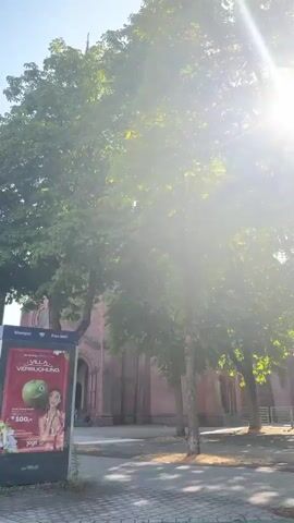 The video opens with a view of a large tree with green leaves, partially obscuring a red brick building with arched windows and a steeple. A digital advertising billboard is visible in the foreground, displaying an advertisement for "Villa Versuchung" with an image of a woman and an apple. The camera pans to the left, revealing more of the red brick building, which appears to be a church. The sun is bright and directly in front of the camera for a moment, causing the image to become overexposed. As the video continues, the camera pans up to show the full facade of the church, including its two spires and clock faces. The sky is blue. There are a few people and bicycles near the entrance of the church. The scene is outdoors, with sunlight and trees suggesting a daytime setting.