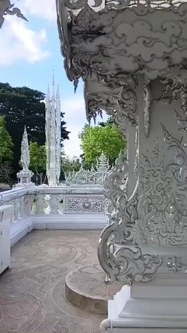 A vertical, eye-level, medium shot captures the ornate white exterior of a temple with intricate, sculpted designs. In the foreground, a white railing adorned with detailed white carvings runs horizontally across the frame. To the left, a white trash can with blue symbols indicating recycling and trash disposal is visible. The background features lush green trees and a bright blue sky with scattered clouds. A tall, slender, pointed spire-like structure made of reflective material stands prominently in the distance, catching the sunlight. The ground in the foreground has a patterned tiled surface. The camera pans slightly to the right, revealing more of the temple's elaborate white architecture and the surrounding greenery.