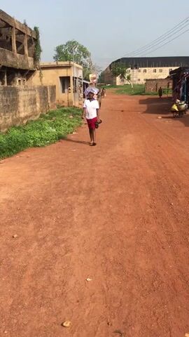 The video features a young woman walking towards the camera on a reddish-brown dirt road. She is dressed in a white t-shirt, a red skirt, and flip-flops, carrying a dark bag in her left hand and a light-colored, possibly wrapped item or shower cap, on her head. The setting is a rural or semi-urban environment in Enyibichiri, Nigeria, characterized by unpaved roads and simple, mostly single-story buildings. Green foliage and bushes line the left side of the road. In the background, other individuals are visible further down the road, including one with what appears to be a wheelbarrow and another near a makeshift stall with hanging clothes. The scene is brightly lit by daylight, suggesting clear weather. There is a street light and power lines visible on the left side of the road. No discernible text is visible in the video.