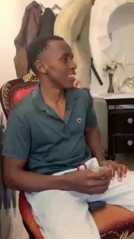 The video shows a room with five people, four of whom are visible in varying degrees. Initially, a young man in a dark green polo shirt and light grey sweatpants sits on a red and gold chair, holding a small, colorful package. Another man in a beige long-sleeved shirt and patterned pants walks behind him. The camera pans slightly to the right, revealing another young man in a light grey sweater and dark jeans, also holding a small, colorful package. Continuing the pan, a third young man in a dark green long-sleeved shirt and tan pants is shown, sitting down. A fourth young man, wearing a light green jacket over a t-shirt and dark pants, walks into the frame and past the camera. In the background, there is a mirror, a small white table with a white vase, and a wardrobe. The people are casually dressed and appear to be relaxed. The room seems to be a living space, possibly an apartment.