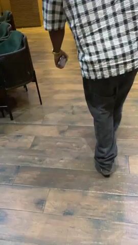 A person is walking across a wooden floor. The person is wearing dark pants, a black and white plaid shirt, and light-colored shoes. The person is holding a phone in their right hand. A chair is partially visible on the left. The floor is made of light brown wooden planks with some blue-grey accents.