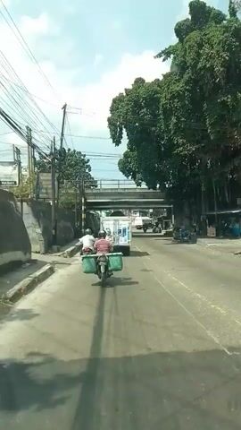 The video shows a view from inside a vehicle traveling on a paved road. In the foreground, a motorcycle with two people on it is moving away from the camera. The motorcycle has two large green containers attached to the back. The driver is wearing a white shirt and a helmet, and the passenger is wearing a striped shirt. On the left side of the road, there is a concrete wall and a sidewalk. On the right side of the road, there are trees and other vehicles, including what appears to be a water truck and a motorcycle. An elevated overpass runs across the road in the background. The sky is visible and appears to be clear. The scene suggests a daytime setting in a possibly urban or semi-urban environment.